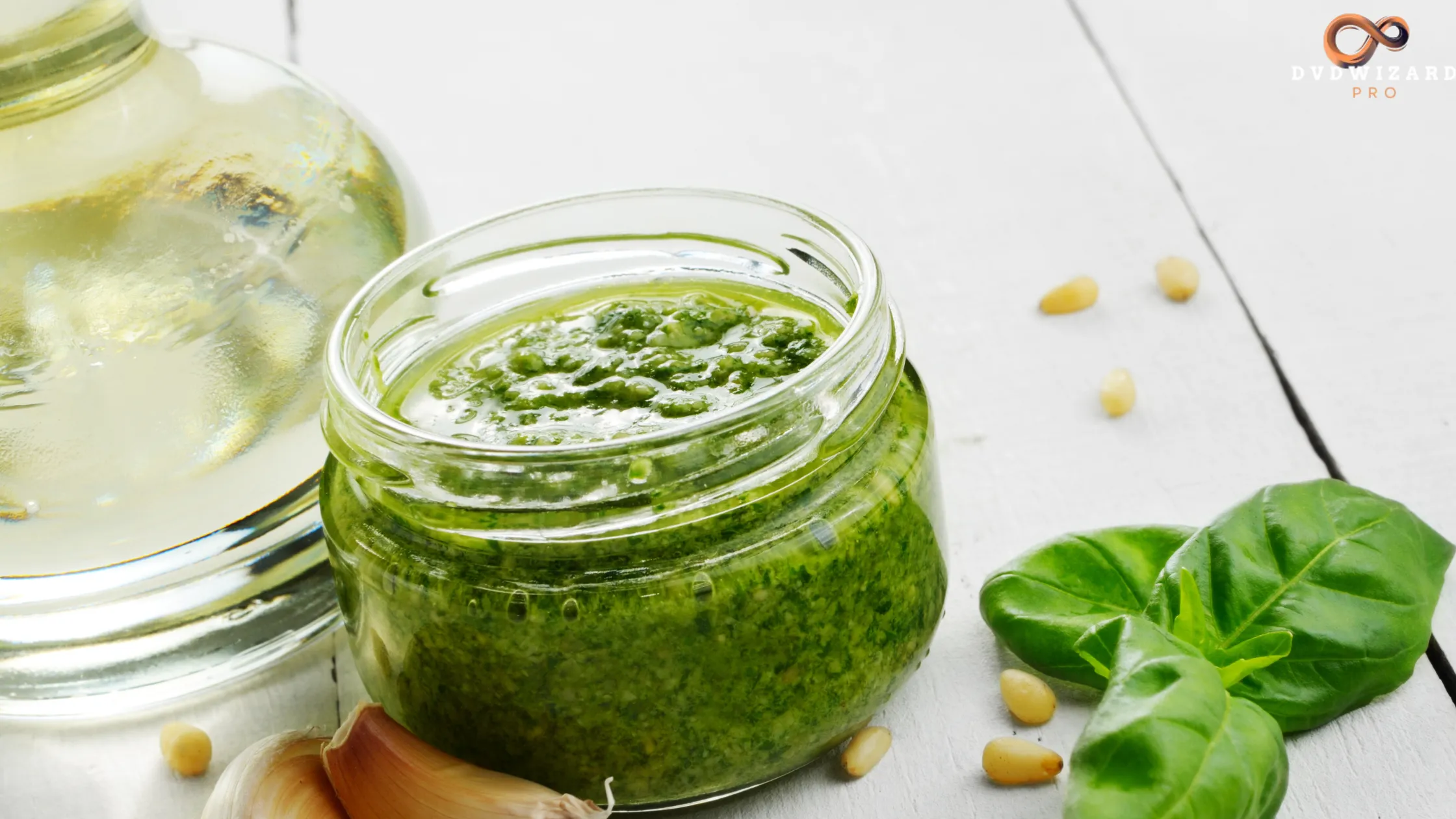 A jar of vibrant Jamaican Green Sauce with fresh basil leaves, pine nuts, garlic, and olive oil on a white wooden surface.